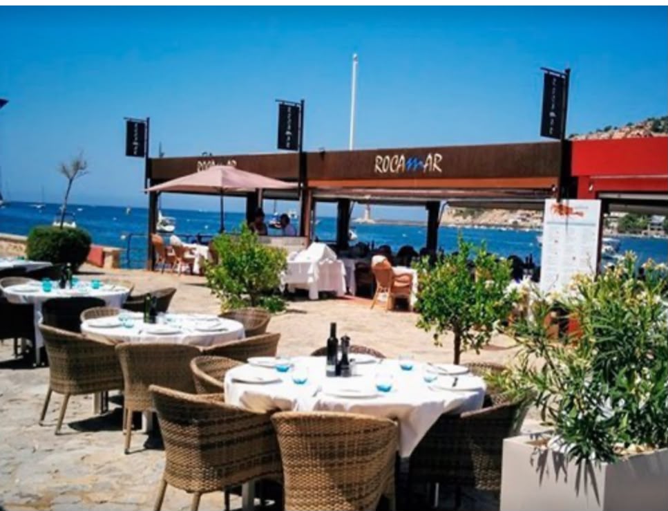 Terraza de Rocamar con mesas vestidas de blanco junto al mar y el cartel del restaurante.